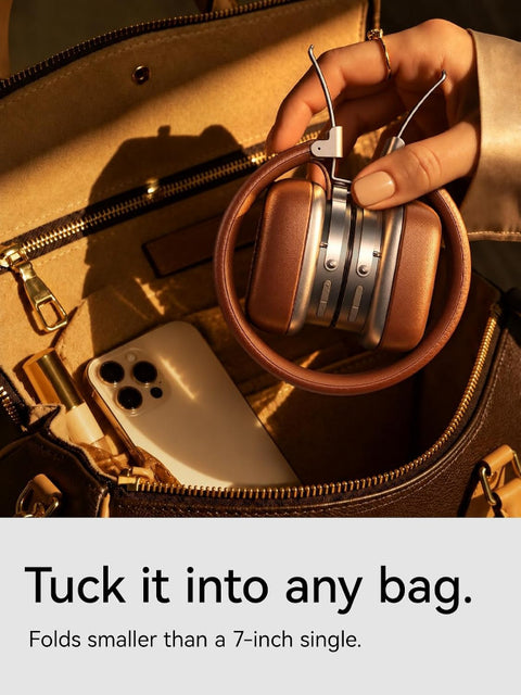 A person holds a compact foldable over-ear headphone with a brown leather-like outer loop and metallic rose-gold earcups, shown tucked inside an open brown handbag. A phone is also visible in the bag.