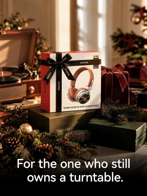 A boxed pair of over-ear headphones sits on a decorated holiday table with pine branches, ornaments, and wrapped gifts in the background. The box is red, white, and black with a ribbon, and the headphones pictured on the packaging appear brown and black.